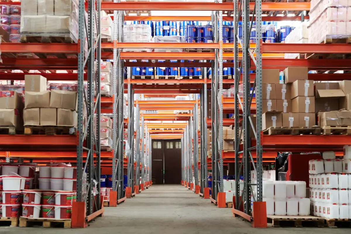 A long warehouse aisle seen down the center, lined with tall orange pallet racks loaded with boxes, drums and pails on pallets