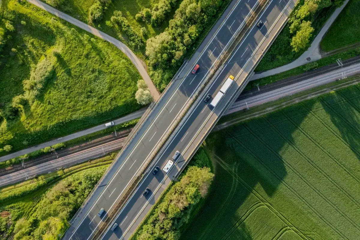 Aerial view of a highway bridge carrying trucks and cars over railway tracks, cutting through green farm fields